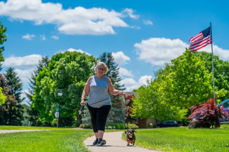 Senior woman walking her dog on a sunny day, with a vibrant American flag and greenery in the background.