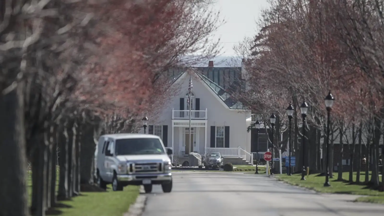 Entrance to white building lined by trees with white van and a few others cars parked in front